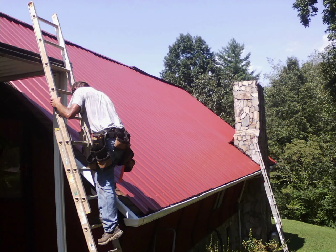 Roofer on a ladder beside a red metal roof and stone chimney — Soffit & Fascia Repair by Roofing Brothers in Missouri Valley